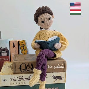 Könnte beinhalten: Eine gehäkelte Puppe in einem gelben Pullover und lila Hosen sitzt auf einem Stapel Bücher. Die Puppe hält ein blaues Buch und hat braunes Haar. Das oberste Buch des Stapels trägt den Text "Horgoláda".