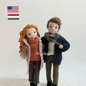 Puede incluir: Dos muñecas de crochet, una niña con el pelo largo y rojo que lleva un jersey marrón y un abrigo beige, y un niño con el pelo marrón que lleva un jersey azul y un abrigo azul marino. Las muñecas están de pie sobre una superficie blanca con puntos blancos.