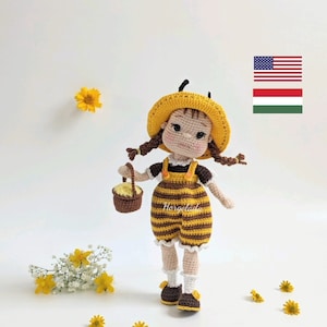 Könnte beinhalten: Eine gehäkelte Puppe, die als Biene verkleidet ist, trägt einen gelben Hut und hält einen kleinen Korb. Die Puppe hat braunes Haar in Zöpfen und steht auf einem weißen Hintergrund mit gelben Blumen.
