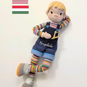 Puede incluir: Una muñeca de crochet con cabello rubio que lleva un mono azul, una camisa a rayas y calcetines a rayas. La muñeca sostiene una honda y tiene una pose relajada. La muñeca está acostada sobre una superficie blanca. El texto "Horgoládda" está bordado en el mono de la muñeca.