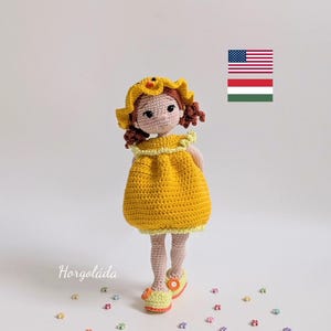 Puede incluir: Muñeca de ganchillo hecha a mano con un vestido amarillo y un sombrero a juego con un detalle floral. La muñeca tiene el pelo castaño y está sobre una superficie blanca. Pequeños adornos con forma de flor de colores están esparcidos.