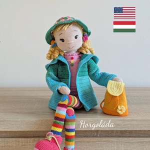 Puede incluir: Muñeca de ganchillo hecha a mano con cabello rubio rizado, que lleva un sombrero y una chaqueta verdes, una camisa rosa y pantalones a rayas arcoíris. La muñeca tiene zapatos rosas y sostiene una pequeña mochila naranja. La muñeca está sentada.