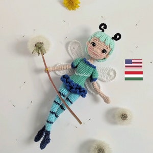 Könnte beinhalten: Eine gehäkelte Feenpuppe mit blauer und grüner Kleidung, weißen Flügeln und schwarzen Antennen. Die Puppe hält einen Löwenzahnstiel. Der Hintergrund ist weiß mit mehreren Löwenzähnen.