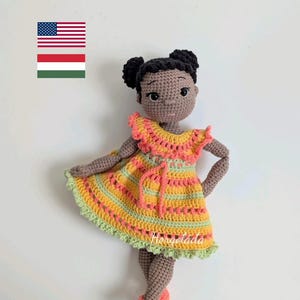 Op de afbeelding: Handgemaakte gehaakte pop met een donkere huid en zwart haar in twee knotjes. De pop draagt een gele, oranje en groene jurk en bijpassende schoenen. De jurk heeft een gerimpelde kraag en een taille met trekkoord.