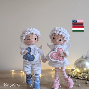 May include: Two handmade crochet angel dolls, one holding a blue heart and the other a pink heart. Both dolls have white curly hair, wings, and are wearing white outfits with blue and pink accents. The dolls are standing on a wooden surface.