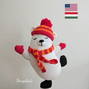 May include: A handmade white crochet polar bear wearing a red and orange striped hat, scarf, and mittens. The bear has black eyes and a black nose. The bear is wearing black boots. The text "Horgoláda" is in the lower left corner.