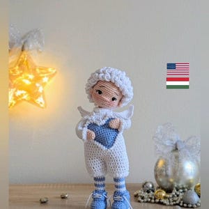 Puede incluir: Muñeca de ángel de ganchillo hecha a mano, blanca con un diseño en forma de corazón azul. La muñeca tiene zapatos azules y calcetines a rayas. Sostiene un corazón azul. La palabra "Horgoláda" está escrita abajo.