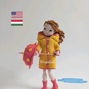 May include: A crocheted doll wearing a yellow raincoat with pink accents and holding a pink and yellow polka dot umbrella. The doll has brown hair and is standing in front of a blue puddle.