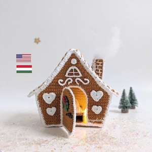 Op de afbeelding: Een gehaakt peperkoekhuis met een witte deur en een krans, verlicht van binnen met een warme gloed. Het huis is versierd met witte hartjes en een witte rand. Het huis staat op een wit oppervlak met witte glitter.