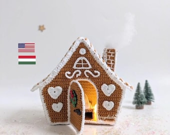 May include: A crocheted gingerbread house with a white door and a wreath, lit from inside with a warm glow. The house is decorated with white hearts and a white trim. The house is sitting on a white surface with white glitter.