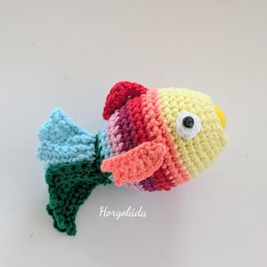 May include: A handmade crochet fish in vibrant colors. The fish features a yellow head, a red dorsal fin, and a multi-colored body with shades of pink, orange, and yellow. The tail is blue and green. The fish has a black button eye and a yellow beak.