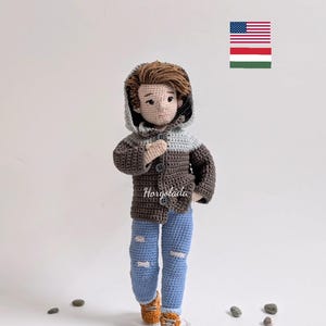 Può includere: Bambola all'uncinetto che indossa una giacca con cappuccio marrone e grigia, jeans blu con dettagli usurati e scarpe arancioni. La bambola ha i capelli castani e un'espressione facciale neutra. La bambola è in piedi su una superficie bianca con piccoli sassi grigi sullo sfondo. L'immagine include anche una piccola bandiera americana e ungherese.