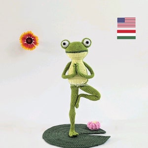Op de afbeelding: Een handgemaakte groene gehaakte kikker in een yogahouding, staand op een groen lelieblad. De kikker heeft grote ogen en houdt zijn handen samen. Links staat een gele en oranje bloem.