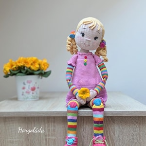 May include: A crocheted doll with blonde hair, a pink dress, rainbow striped sleeves and legs, and pink shoes. The doll is holding a yellow flower and sitting on a wooden surface.