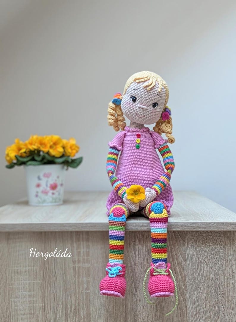 May include: A crocheted doll with blonde hair, a pink dress, rainbow striped sleeves and legs, and pink shoes. The doll is holding a yellow flower and sitting on a wooden surface.