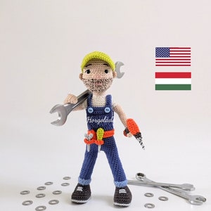 Può includere: Giocattolo amigurumi all'uncinetto di un operaio che indossa una tuta blu, un casco giallo e tiene una chiave inglese. Il giocattolo è in piedi su una superficie bianca con diversi lamierini metallici sparsi intorno. Il giocattolo tiene un trapano nella sua mano destra. Il testo "Horgoládá" è visibile sulla tuta del giocattolo.