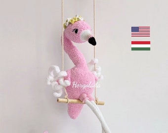 May include: Pink crocheted flamingo doll with white wings and green shoes, sitting on a wooden swing. The doll is wearing a white flower crown and has black eyes. The background is white with pink and green flowers.