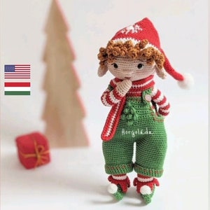 May include: A crocheted Christmas elf doll wearing a red and white striped sweater, green overalls, and a red hat with a white pom-pom. The elf is holding its hands together in front of its chest. The doll is standing in front of a wooden Christmas tree and a red gift box.