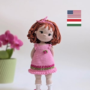 Könnte beinhalten: Handgefertigte Häkelpuppe mit braunen Locken, die ein rosa Kleid mit floralem Besatz und passenden Schuhen trägt. Die Puppe hat ein rosa Stirnband und steht vor einem weißen Hintergrund. Die Puppe ist etwa 20 cm groß.
