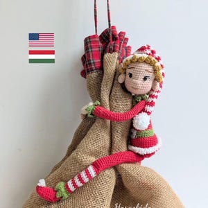 Puede incluir: Una muñeca de elfo navideño hecha a mano, tejida con brazos y piernas a rayas rojas y blancas, un gorro a rayas rojas y blancas y un cuello a rayas verdes y blancas. La muñeca abraza una bolsa de arpillera con una parte superior a cuadros rojos.