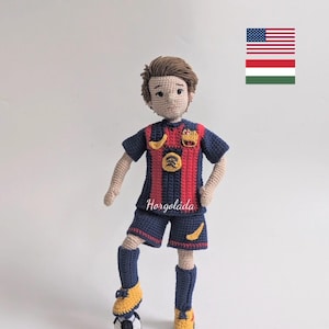 Può includere: Bambola di calciatore all'uncinetto con maglia a righe blu e rosse, pantaloncini blu e scarpe gialle e blu. La bambola è in piedi su un pallone da calcio. Il testo "Horgoláda" è visibile.