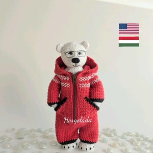 May include: A white crocheted polar bear wearing red and white striped overalls with a black zipper and a black hooded jacket. The bear is wearing black framed glasses and has a black nose and mouth. The bear is standing on a white surface with white stones.