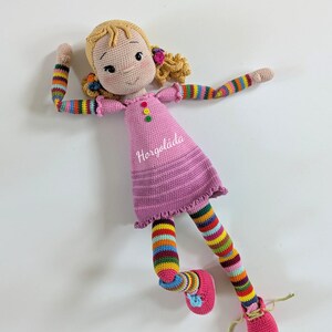 May include: A crocheted doll with blonde hair, a pink dress, colourful striped sleeves and leggings, and pink shoes. The doll is lying on its back with its arms and legs outstretched.