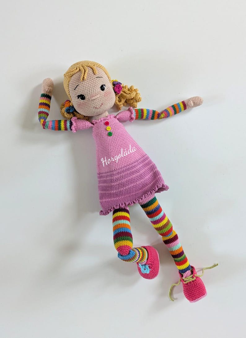 May include: A crocheted doll with blonde hair, a pink dress, colourful striped sleeves and leggings, and pink shoes. The doll is lying on its back with its arms and legs outstretched.