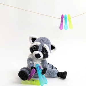 K&ouml;nnte beinhalten: Ein graues und wei&szlig;es geh&auml;keltes Pl&uuml;sch-Waschb&auml;r-Spielzeug, das auf einer wei&szlig;en Oberfl&auml;che sitzt. Der Waschb&auml;r h&auml;lt eine lila und blaue W&auml;scheklammer. &Uuml;ber dem Waschb&auml;r h&auml;ngen drei W&auml;scheklammern an einer Schnur.