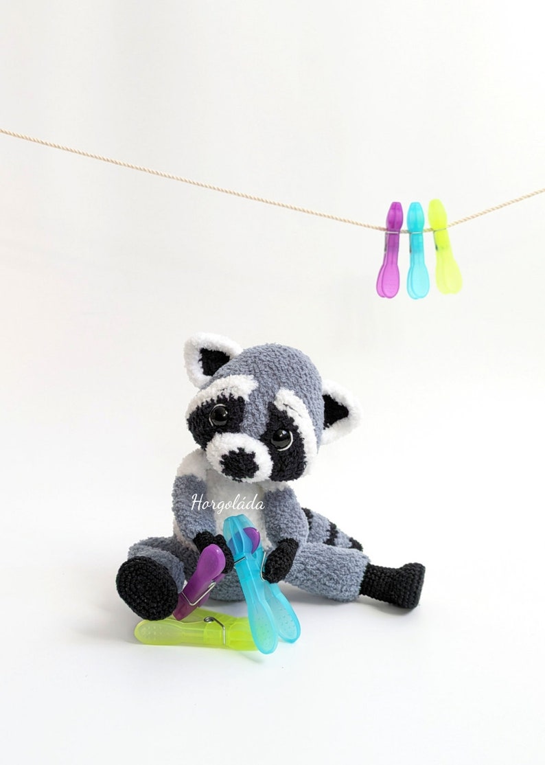K&ouml;nnte beinhalten: Ein graues und wei&szlig;es geh&auml;keltes Pl&uuml;sch-Waschb&auml;r-Spielzeug, das auf einer wei&szlig;en Oberfl&auml;che sitzt. Der Waschb&auml;r h&auml;lt eine lila und blaue W&auml;scheklammer. &Uuml;ber dem Waschb&auml;r h&auml;ngen drei W&auml;scheklammern an einer Schnur.
