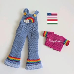 May include: A light blue crocheted overall with two yellow smiley face buttons, a rainbow and cloud applique, and rainbow stripes at the cuffs. Beside it is a magenta crocheted top with rainbow striped sleeves and the text Horgoláda.