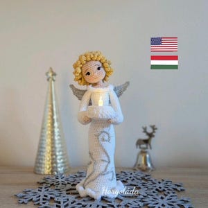 Könnte beinhalten: Eine gehäkelte Engelfigur, die eine Kerze hält, steht auf einer Schneeflockenmatte. Der Engel hat blonde Haare, ein weißes Kleid und silberne Akzente. Die Figur ist von Schneeflocken und einem silbernen Weihnachtsbaum umgeben.