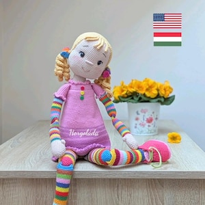 Może przedstawiać: Lalka szydełkowa z blond włosami, różową sukienką i kolorowymi, paskowanymi nogami i rękami. Lalka siedzi na drewnianej powierzchni z doniczką żółtych kwiatów w tle. Tekst "Horgolata" jest widoczny na sukience lalki.