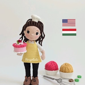 Puede incluir: Muñeca de crochet con gorro de cocinero y delantal, sosteniendo un pastel rosa y blanco. La muñeca tiene el pelo castaño y lleva pantalones negros y zapatos marrones. Hay dos ovillos de lana, un batidor y un molde para cupcakes en el fondo. El texto "Horgoláda" es visible en el delantal de la muñeca.