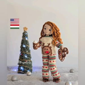 Puede incluir: Muñeca de crochet con un pijama de temática de reno con una taza de chocolate caliente en una mano y un juguete con forma de reno en la otra. La muñeca está de pie frente a un pequeño árbol de Navidad con luces blancas.