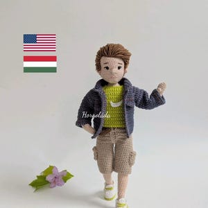 以下が含まれることがあります： 緑色のシャツ、カーキ色のショートパンツ、青いジャケットを着た人の、かぎ針編みの人形。人形は茶色の髪と白いスニーカーをしています。人形は前方に歩いています。「Horgolada」の文字がジャケットにあります。