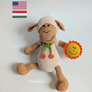 May include: A handmade crochet sheep doll with a smiling face, wearing a green scarf with orange flowers, and holding a yellow sun. The doll is beige, brown, and white. The word "Horgoláda" is written on the bottom.