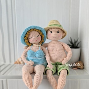 Puede incluir: Dos muñecos de ganchillo hechos a mano, una mujer y un hombre, están sentados juntos. La mujer lleva un bañador y un sombrero azules, mientras que el hombre lleva pantalones cortos verdes y un sombrero de paja. Los muñecos están hechos de hilo y tienen los ojos cerrados.