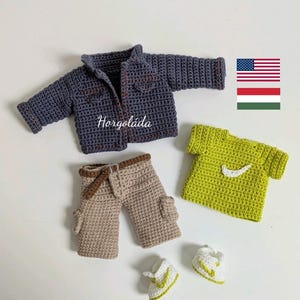 May include: A collection of crocheted doll clothes in blue, beige, and green. The set features a jacket, trousers, a t-shirt with a white design, and tiny trainers. The jacket and trousers have brown detailing. The t-shirt is lime green.