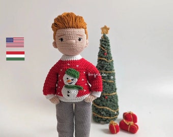 Puede incluir: Muñeca de crochet con un suéter rojo con un diseño de muñeco de nieve, pantalones grises y zapatos negros. La muñeca está de pie frente a un pequeño árbol de Navidad verde con luces doradas y adornos rojos y dorados.