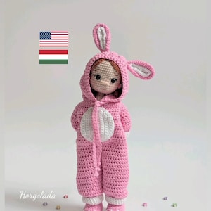 May include: A handmade crochet doll wearing a pink bunny costume with a hood and long ears. The doll has a white belly and white accents on the feet. The doll is standing on a white surface with small colorful flower-shaped decorations.