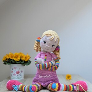 May include: A crocheted doll with blonde hair, pink dress, and rainbow striped arms and legs. The doll is sitting on a wooden surface with a flower pot in the background.
