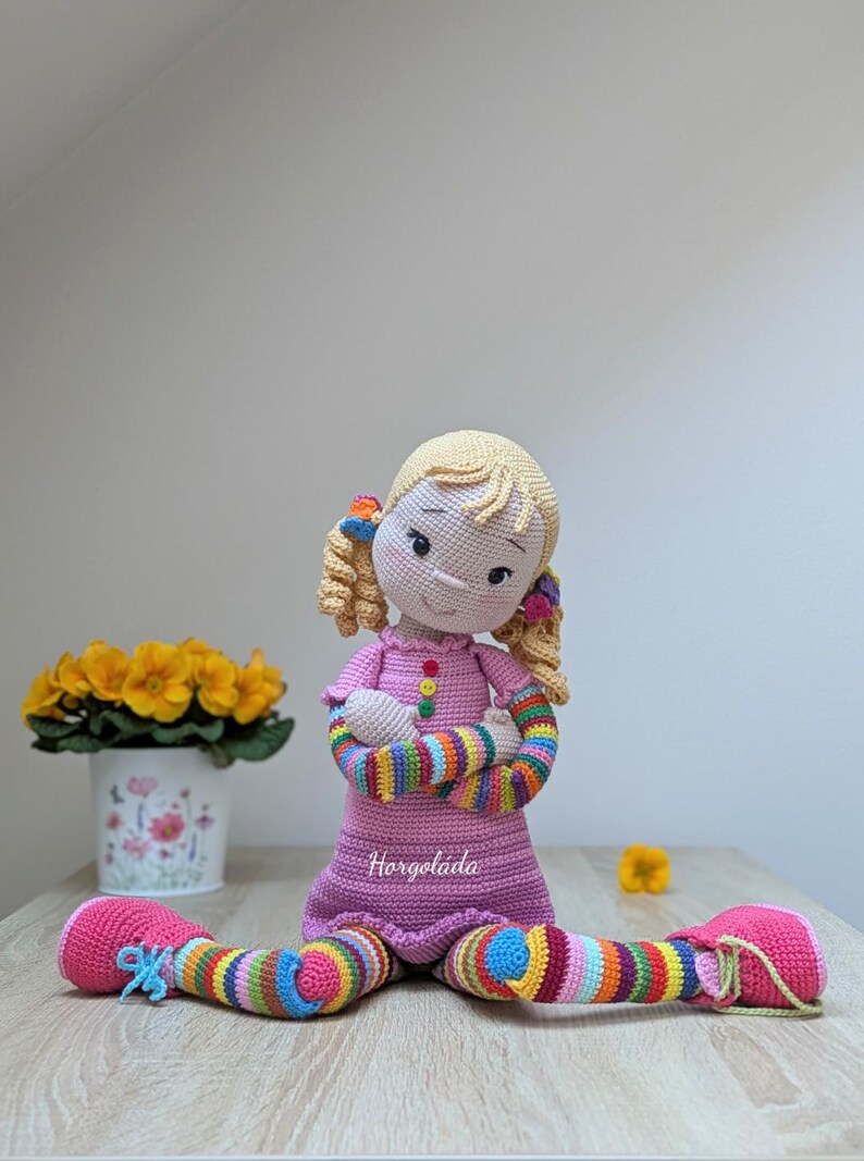 May include: A crocheted doll with blonde hair, pink dress, and rainbow striped arms and legs. The doll is sitting on a wooden surface with a flower pot in the background.