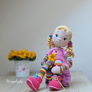 May include: A crocheted doll with blonde hair, pink dress, rainbow striped sleeves and leggings, and pink shoes. The doll is sitting on a wooden surface.
