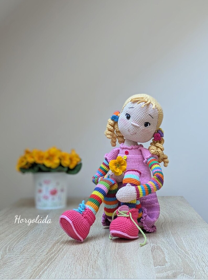 May include: A crocheted doll with blonde hair, pink dress, rainbow striped sleeves and leggings, and pink shoes. The doll is sitting on a wooden surface.