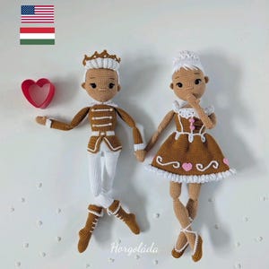 May include: Two crocheted dolls, a prince and a ballerina, stand side-by-side. The prince wears a brown and white uniform with a crown. The ballerina wears a brown dress with pink hearts and ballet shoes. A red heart-shaped cookie cutter is on the left.
