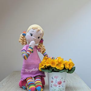 May include: A crocheted doll with blonde hair and a pink dress sits next to a white pot with yellow flowers. The doll's arms and legs are striped with rainbow colours. The pot has a floral design.