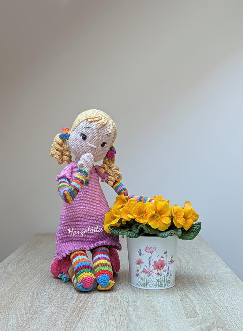 May include: A crocheted doll with blonde hair and a pink dress sits next to a white pot with yellow flowers. The doll's arms and legs are striped with rainbow colours. The pot has a floral design.