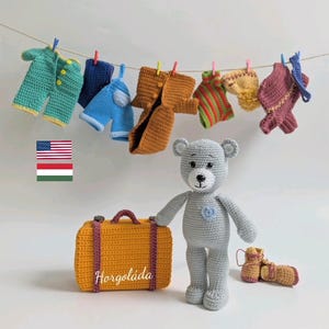 May include: A gray crocheted teddy bear with a blue heart on its chest stands next to a yellow crocheted suitcase with the word "Horgoláda" on it. The bear is wearing brown crocheted boots. There are seven crocheted outfits hanging on a clothesline behind the bear.