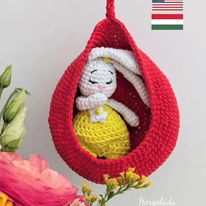 May include: A handmade crochet bunny doll in a red hanging pod. The bunny has white fur, long ears, and a yellow dress. The pod is suspended by a red cord. The word "Horgoláda" is visible in the image.